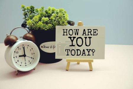 How are you today? written on paper card with artificial plant and alarm clock on blue background