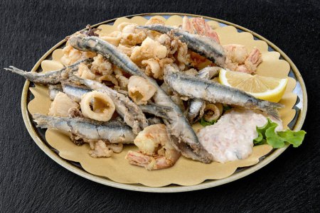 Round dish with mixed fried fish with sauce and lemon wedge on black stone 