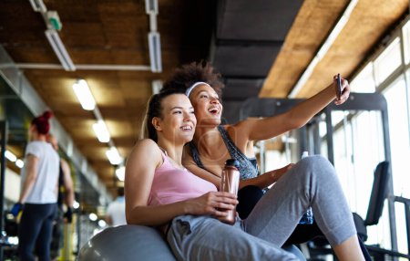 Photo for Happy fit women, friends smiling, talking and taking photos after work out in gym. Social media, people, sport concept. - Royalty Free Image