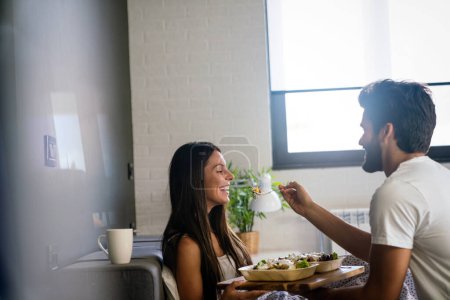 Téléchargez les photos : Beau jeune homme romantique apporte le petit déjeuner à la femme dans le lit et rire ensemble après le réveil. - en image libre de droit