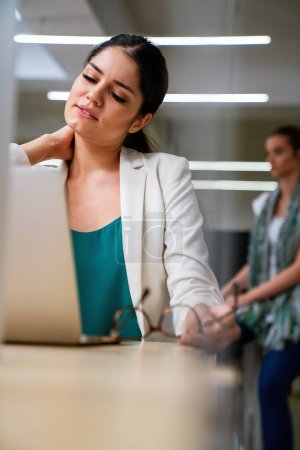 Foto de Young attractive woman at modern office desk, working on laptop, with headaches, noisy loud office giving a migraine, relieving stress, chronic pain. - Imagen libre de derechos