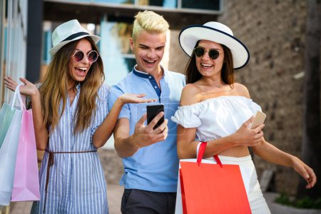 Téléchargez les photos : Groupe d'amis heureux utilisant le téléphone mobile pendant les achats en plein air - en image libre de droit