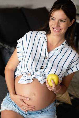 Foto de Embarazo, descanso, personas y concepto de expectativa. Feliz hermosa embarazada tocando su vientre en casa - Imagen libre de derechos