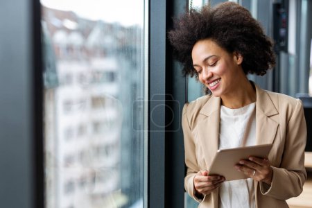 Téléchargez les photos : Portrait de jeune femme afro-américaine réussie travaillant avec une tablette au bureau - en image libre de droit