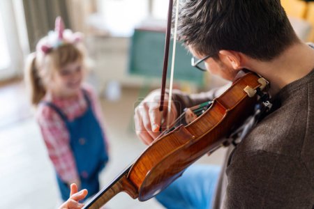 Photo for Charming little girl learning to play the violin with an artistic music teacher. Young man teaching a kid student at home - Royalty Free Image