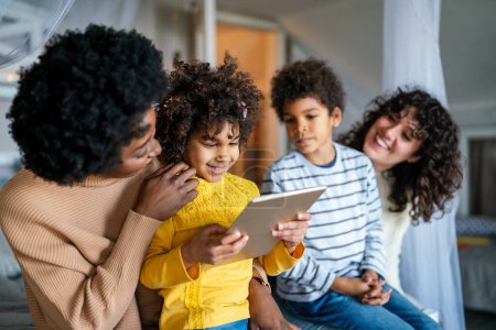 Foto de Familia multiétnica feliz divirtiéndose con dispositivo digital en casa, padres homosexuales y niños pequeños usando tableta digital juntos. - Imagen libre de derechos