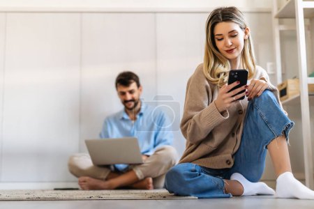 Foto de Mujer joven sonriente usando el teléfono inteligente con el hombre sentado con el ordenador portátil. La gente trabaja en redes sociales concepto de oficina en casa. - Imagen libre de derechos