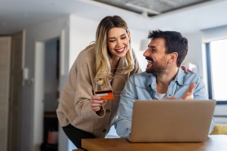 Foto de Alegre joven pareja utilizando el ordenador portátil y sonriendo mientras que las compras en línea en casa juntos - Imagen libre de derechos