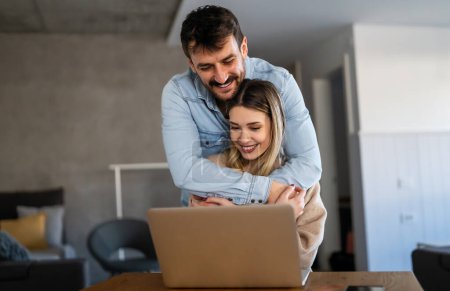Téléchargez les photos : Joyeux jeune couple utilisant un ordinateur portable et souriant tout en faisant du shopping en ligne à la maison ensemble - en image libre de droit