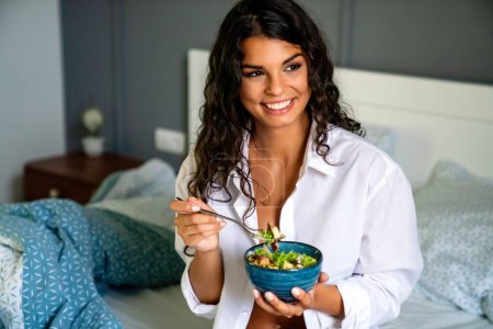 Foto de Retrato de una joven y sexy mujer feliz comiendo ensalada saludable en el dormitorio. Concepto de dieta saludable. - Imagen libre de derechos