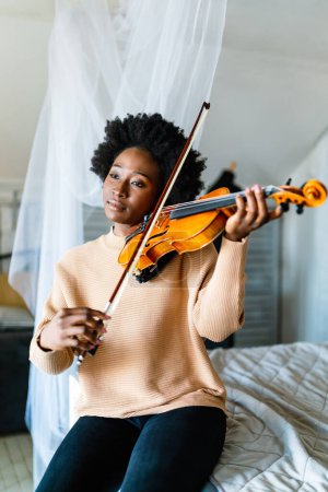 Photo for Attractive black young woman musician plays the violin practicing musical instrument at home - Royalty Free Image