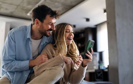 Téléchargez les photos : Jeune couple heureux utilisant le téléphone pour partager les nouvelles des médias sociaux à la maison, souriant parler faire du shopping en ligne. Concept de mode de vie mobile tech - en image libre de droit