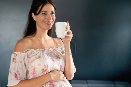 Téléchargez les photos : Belle jeune femme enceinte heureuse souriante et buvant café, thé ou vitamines à la maison - en image libre de droit