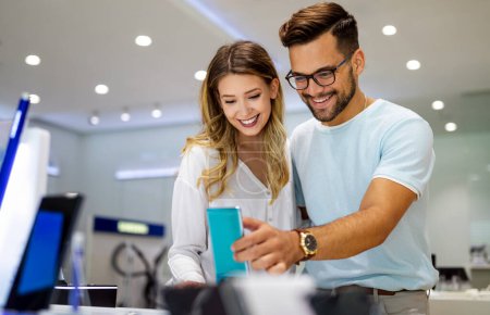 Photo for Young satisfied couple testing the new model of a mobile phone from the desk in the tech store. - Royalty Free Image