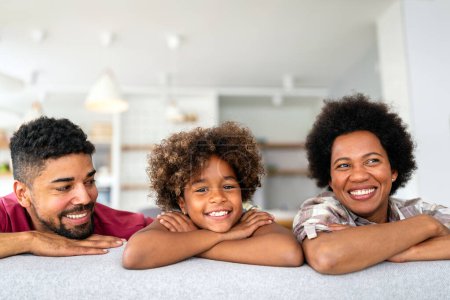 Téléchargez les photos : Portrait d'une jeune famille afro-américaine souriante avec une petite fille adolescente assise sur un canapé à la maison profiter du week-end ensemble. - en image libre de droit