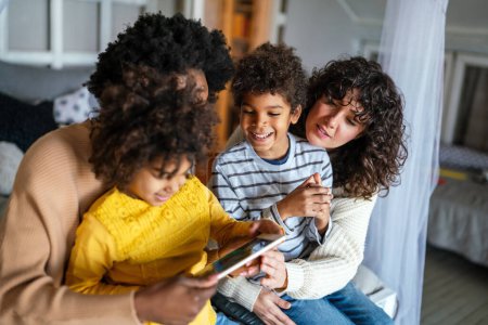 Téléchargez les photos : Heureuse famille multiethnique s'amuser avec un appareil numérique à la maison, les parents homosexuels et les petits enfants utilisant la tablette numérique ensemble. - en image libre de droit
