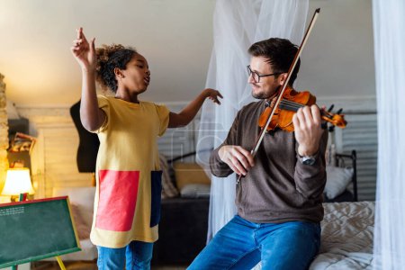 Téléchargez les photos : La musique est tellement amusante. Le jeune père apprend à sa petite fille adoptive à jouer du violon et à sourire. Concept de bonheur des enfants monoparentaux. - en image libre de droit