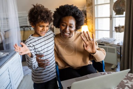 Téléchargez les photos : Heureuse mère célibataire afro-américaine et son fils saluant sur appel vidéo par ordinateur portable à la maison. Technologie gens concept de communication en ligne - en image libre de droit