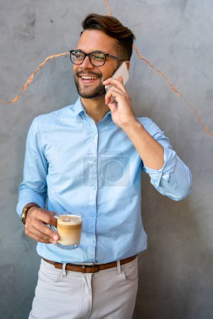 Photo for Portrait of a smiling young handsome business man using smartphone and holding cup of coffee - Royalty Free Image