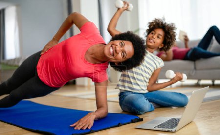 Foto de Feliz joven afroamericana mujer con niño viendo en línea video entrenamiento deportivo y estiramiento en la estera de fitness. Concepto de familia, salud, deporte y yoga en casa - Imagen libre de derechos