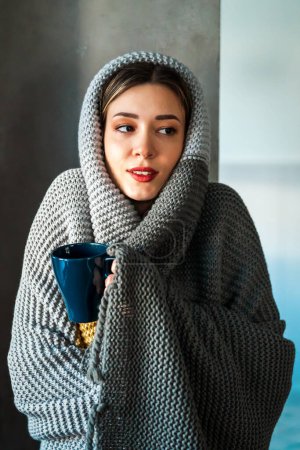 Photo for Low temperature in the apartment, no heating. Tired and sick woman is covered with a blanket, holding a cup of hot drink, medicine. Suffers from pain, weakness. - Royalty Free Image