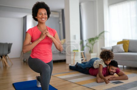 Foto de Mujer afroamericana, madre haciendo ejercicio en casa en la sala de estar, padre jugando con un niño de fondo. Concepto familiar de deporte doméstico - Imagen libre de derechos