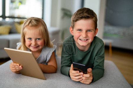 Téléchargez les photos : Portrait de petits enfants intelligents tenant smartphone jouant jeu mobile en ligne à la maison, concept enfant et gadget - en image libre de droit