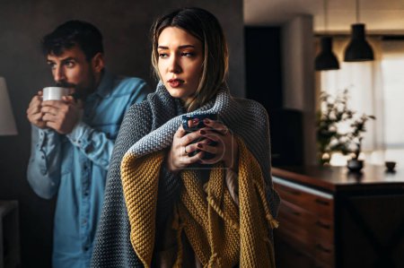 Photo for Sad sick young couple wrapped in blanket drinking hot tea trying to warm up. Freezing man and woman suffering from cold or flu fever or having trouble with central heating - Royalty Free Image