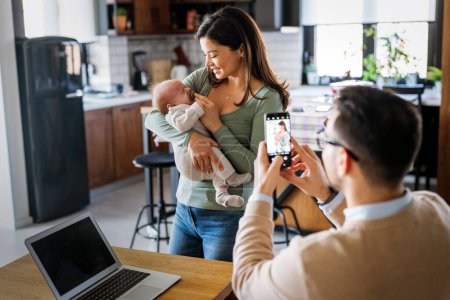 Foto de Orgulloso padre joven tomar una foto de su esposa con bebé en casa. Familia feliz, concepto tecnológico. - Imagen libre de derechos