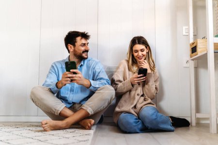 Téléchargez les photos : Couple souriant regardant dans le smartphone ensemble à la maison. Les jeunes gens heureux utilisant deux téléphones partagent les nouvelles des médias sociaux à la maison - en image libre de droit