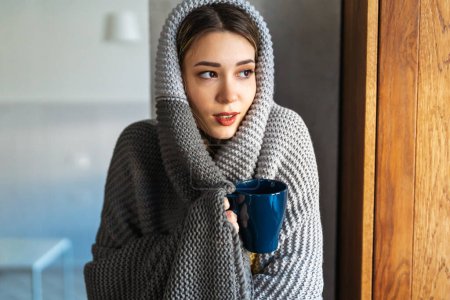 Photo for Low temperature in the apartment, no heating. Tired and sick woman is covered with a blanket, holding a cup of hot drink, medicine. Suffers from pain, weakness. - Royalty Free Image