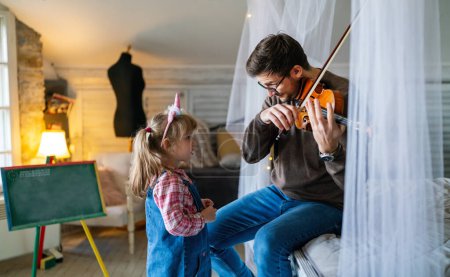 Foto de Encantadora niña aprendiendo a tocar el violín con un profesor de música artística. Joven enseñando a un niño estudiante en casa - Imagen libre de derechos