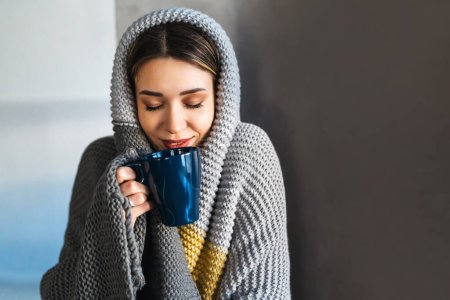 Photo for Happy young woman at home in winter wrapped herself in a warm cozy blanket, holding a cup of hot drink - Royalty Free Image
