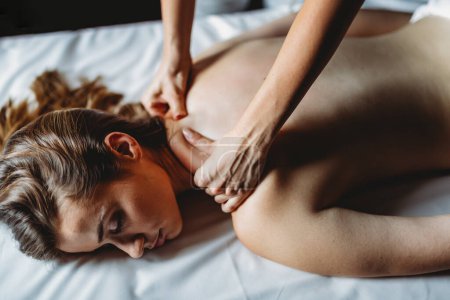 Téléchargez les photos : Joyeuse masseuse professionnelle masseuse dos de jeune cliente détendue sur table de massage dans un salon de spa - en image libre de droit