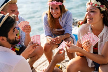 Photo for Group of young smiling friends having fun while playing cards on beach at summer vacation - Royalty Free Image