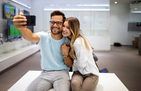 Téléchargez les photos : Beau jeune couple est câlin, faire un selfie à l'aide d'un téléphone intelligent et souriant. Appareil numérique, concept d'intelligence artificielle. - en image libre de droit