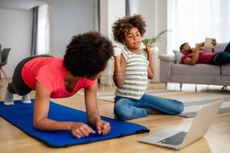 Téléchargez les photos : Joyeux jeune mère et fille pratiquant le yoga en ligne, ensemble à la maison, assis sur un tapis de fitness, jouissant d'un mode de vie sain - en image libre de droit