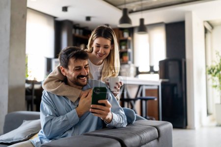 Photo for Happy young couple, woman and man hugging, using smartphone together. Smiling overjoyed wife and husband looking at phone screen. Social media concept. - Royalty Free Image