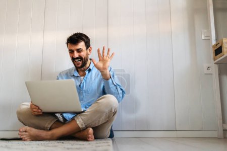Téléchargez les photos : Un bel homme heureux utilisant un ordinateur portable fait du travail à distance. Homme assis sur le sol utilise Internet, écrit rapport de projet. Travailleur éloigné efficace et productif de la maison. - en image libre de droit
