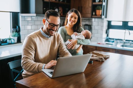 Photo for Young father works on laptop while his wife looks after child. Work from home, parenting, telecommuting - Royalty Free Image