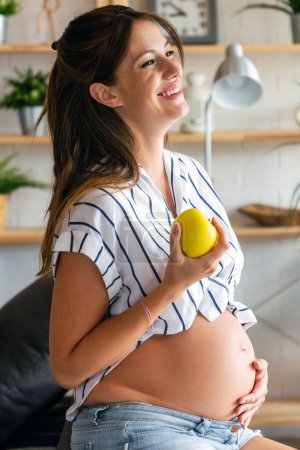Téléchargez les photos : Grossesse, repos, personnes et concept d'attente. Heureuse belle femme enceinte touchant son ventre à la maison - en image libre de droit