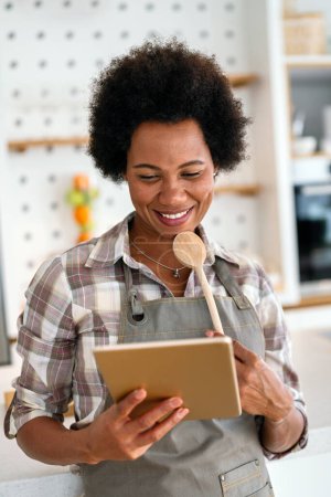 Téléchargez les photos : Jeune femme afro-américaine utilisant une tablette pour cuisiner dans sa cuisine. Technologie des personnes concept alimentaire. - en image libre de droit