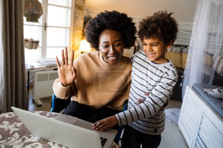Foto de Feliz afroamericana madre soltera con el niño saludando las manos mientras hace videollamadas a través de la computadora portátil en casa. Gente, concepto de comunicación de tecnología familiar - Imagen libre de derechos