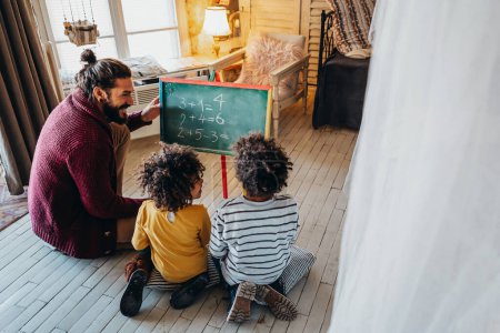 Téléchargez les photos : Enfants heureux d'apprendre les mathématiques avec un père adoptif diversifié, enseignant à la maison. Éducation familiale concept bonheur - en image libre de droit