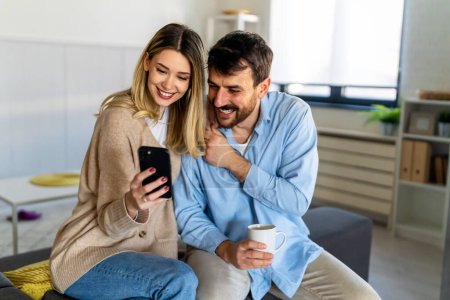 Young happy couple using phone to share social media news at home, smiling talking doing shopping online. Mobile tech lifestyle concept