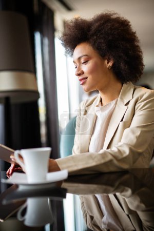 Foto de Atractivo éxito joven afroamericana mujer de negocios beber un café y el uso de una tableta digital durante un descanso - Imagen libre de derechos