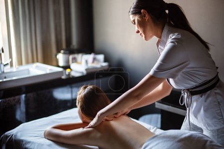 Téléchargez les photos : Joyeuse masseuse professionnelle masseuse dos de jeune cliente détendue sur table de massage dans un salon de spa - en image libre de droit