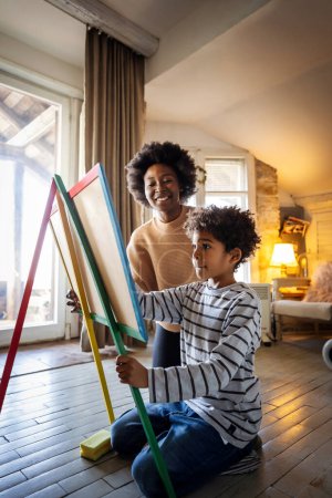Madre soltera afroamericana con hijo dibujando a bordo con tiza juntos. Mujer y niño preescolar divirtiéndose en casa, familia involucrada en actividades creativas
