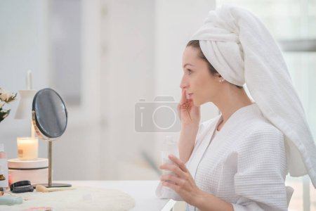 Téléchargez les photos : Belle femme en peignoir et avec une serviette sur la tête qui nettoie son visage avec une éponge de coton. routine beauté, concept d'autosoin. - en image libre de droit