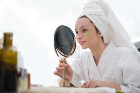 Téléchargez les photos : Beautiful woman in bathrobe and  towel wrapped on head applying eye patches in front of the mirror. - en image libre de droit